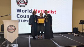 Drei Personen stehen auf einer Bühne vor einer großen Leinwand mit der Aufschrift 'World Methodist Council El Salvador'. Zwei Personen tragen lange, dunkle Talare, die mittlere Person (Rosemarie Wenner) hält eine gerahmte Urkunde und trägt Alltagskleidung. Alle drei lächeln. Links steht ein Rednerpult mit einem weißen Tuch, auf dem das Logo des World Methodist Council zu sehen ist. Im Hintergrund rechts steht ein Notenständer mit Mikrofon.