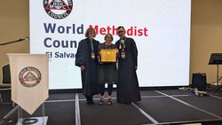 Drei Personen stehen auf einer Bühne vor einer großen Leinwand mit der Aufschrift 'World Methodist Council El Salvador'. Zwei Personen tragen lange, dunkle Talare, die mittlere Person (Rosemarie Wenner) hält eine gerahmte Urkunde und trägt Alltagskleidung. Alle drei lächeln. Links steht ein Rednerpult mit einem weißen Tuch, auf dem das Logo des World Methodist Council zu sehen ist. Im Hintergrund rechts steht ein Notenständer mit Mikrofon.