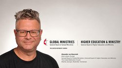 Links ein Mann mittleren Alters mit gegelten Haaren und Ohrring links sieht durch eine Brille freundlich direkt in die Kamera. Rechts das Logo der E m K und die Namen zweier Generalbehörden. Darunter der Name des Mannes und seine neue Stellenbezeichnung.