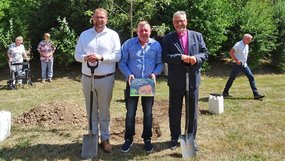 Christoph Ihling, Rainer König und Bischof Werner Philipp stehen vor einem jungen Apfelbaum. Ihling und Philipp halten Spaten, König zeigt ein Schild zur Baumpflanzung; im Hintergrund sind weitere Teilnehmende zu sehen.