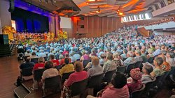 Eine große Halle mit vielen Gottesdienst-Besuchern. Auf der Bühne stehen eine Frau und ein Mann, die die Besucher ansprechen. Hinter ihnen sitzt der Posaunenchor. Die Bühne ist mit Blumen geschmückt.