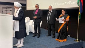 Seitlicher Blick auf eine Bühne: Ein Mann mit Turban spricht an einem Stehpult in zwei Mikrofone. Dahinter steht Bischof Rückert in Kollarhemd und dunklem Anzug mit Bischofsnadel am Revers. Dahinter ein Mann mit Kippa. Daneben eine Frau in schwarz-orangefarbenem Kleid.