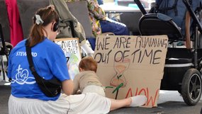Eine junge Frau in einem weißen Rock und blauen T-Shirt sitzt auf dem Boden im Sitzungssaal. Vor ihr stehen zwei selbst gemalte Pappschilder mit der Aufschrift „We are running out of time” und „Save the planet”.