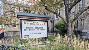 Vor dem steinernen Kirchengebäude steht unter einem Baum eine große Willkommenstafel der Knox United Church in Calgary. Darauf steht auf Englisch: »Welcome worldwide UMC bishops and guests!«
