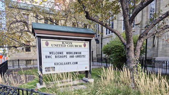 Vor dem steinernen Kirchengebäude steht unter einem Baum eine große Willkommenstafel der Knox United Church in Calgary. Darauf steht auf Englisch: »Welcome worldwide UMC bishops and guests!«