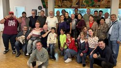Rund dreißig Personen unterschiedlichen Alters haben sich in einem großen Raum zu einem Gruppenfoto versammelt. Darunter sind auch zwei Kinder. Eine Uhr im Hintergrund zeigt kurz vor halb eins.