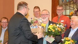 Ein Mann in grauem Jackett übergibt einem anderen Mann ebenfalls in einem grauen Jackett ein Paket mit Geschenken. Darum herum stehen weitere vier Männer, teils mit Blumen in der Hand. Sie lächeln.