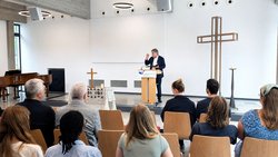 In einem größeren Raum sitzen im Vordergrund Menschen auf Holzstühlen. Sie sehen auf einen Mann mit graumelierten Haaren, Brille, offenem Hemd, schwarzem Anzug, der an einem Pult steht und spricht. In einer Hand hält er ein Buch, mit der anderen gestikuliert er.