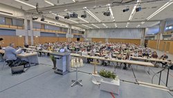 Großer Saal mit vielen Menschen, die an langen Tischen sitzen und nach vorne blicken. Im Vordergrund stehen ein Rednerpult mit Mikrofonen, mehrere Kerzen auf einem Tisch, ein Holzkreuz und Blumenschmuck. Die Decke ist mit zahlreichen Leuchten und technischen Installationen ausgestattet. Die Wände sind hell und mit Holzpaneelen verkleidet, große Fenster lassen Tageslicht herein.