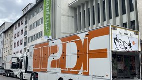 Ein großer weiß-orangefarbener ZDF-Übertragungswagen steht vor der Hoffnungskirche in Stuttgart. Am geöffneten Heck sind zahlreiche technische Geräte und Kabel zu sehen. Im Hintergrund hängt eine grüne Fahne mit der Aufschrift »Hoffnungskirche«.