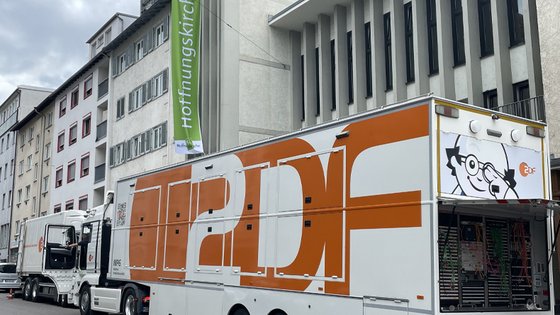 Ein großer weiß-orangefarbener ZDF-Übertragungswagen steht vor der Hoffnungskirche in Stuttgart. Am geöffneten Heck sind zahlreiche technische Geräte und Kabel zu sehen. Im Hintergrund hängt eine grüne Fahne mit der Aufschrift »Hoffnungskirche«.