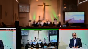 Im Vordergrund sind zwei Bildschirme. Sie zeigen in Programmfenstern einige Kameraperspektiven wie den Gottesdienstraum mit einigen Konferenzteilnehmern an Tischen und den Bischof am Rednerpult. Der unscharfe Hintergrund ist der Gottesdienstraum.