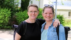 Die Frau links, kurzes blondes Haar, Brille, dunkles T-Shirt, Rucksack, legt der Frau rechts, nach hinten frisiertes blondes Haar, Brille im Haar, Bluse und Jeansjacke, ebenfalls mit Rucksack, ihren linken Arm um den Hals. Beide lächeln in die Kamera.