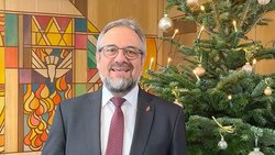 Das Startbild der Video-Weihnachtsbotschaft zeigt Bischof Harald Rückert. Er trägt ein graues Jackett, ein weißes Hemd und eine rote Krawatte. Hinter ihm ist rechts der geschmückte Weihnachtsbaum und links die bunt gestaltete Wand eines Altarraums.