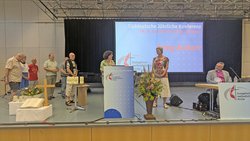 Auf einer Bühne: In der Mitte steht an einem Stehpult eine Frau und spricht eine Frau rechts daneben an. Ganz rechts sitzt der Bischof an einem Tisch und sieht zu. Ganz links stehen sechs weitere Personen und sehen größtenteils zu.