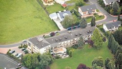 Schräg von oben aus Nordwesten das langgestreckte Anwesen von Haus Höhenblick. Im Norden schließt sich ein Parkplatz an. Am Anwesen führt im Osten eine Straße entlang. Im Westen befinden sich ein Rasen sowie Bäume und Gebüsch.