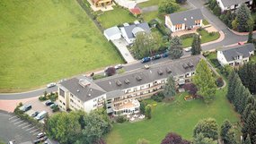 Schräg von oben aus Nordwesten das langgestreckte Anwesen von Haus Höhenblick. Im Norden schließt sich ein Parkplatz an. Am Anwesen führt im Osten eine Straße entlang. Im Westen befinden sich ein Rasen sowie Bäume und Gebüsch.