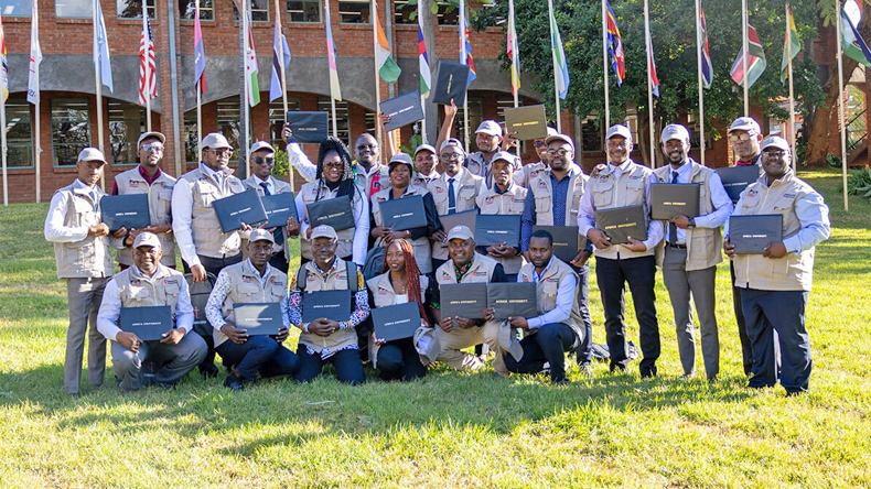 Die erste der beiden Gruppen nach der Abschlussfeier am 26. Juni an der Africa University in Mutare. Die Weiterbildung stärkt die Öffentlichkeitsarbeit der EmK bei Krisen, Krankheitsausbrüchen und Falschmeldungen. Mehr als zwanzig Teilnehmende stehen und knien mit ihren Urkunden auf einer Wiese vor der Africa University. Sie tragen helle Westen und Kappen; im Hintergrund wehen zahlreiche Nationalflaggen.