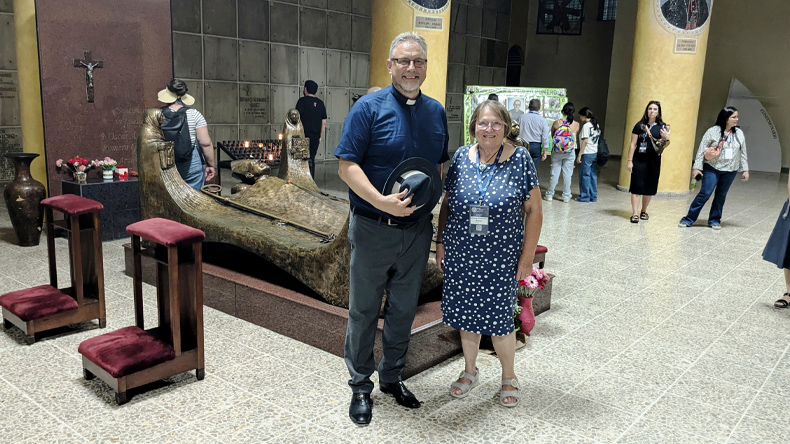 Der Besuch am Grab Óscar Romeros führt die Tagungsteilnehmenden an einen zentralen Erinnerungsort El Salvadors. Im Bild: Bischof Werner Philipp und Bischöfin i. R. Rosemarie Wenner. Im Mittelpunkt Bischof Werner PHilipp in dunkler Kleidung mit weißem Kollar, der neben Bischöfin in Ruhe Rosemarie Wenner im gepunkteten Kleid steht. Beide lächeln in die Kamera. Sie befinden sich in einer Halle mit gelbem Säulen, Mosaikfenstern und einer großen bronzenen Skulptur in Form eines liegenden Körpers. Im Hintergrund sind weitere Menschen zu sehen, einige fotografieren oder betrachten die Umgebung. Links im Bild stehen zwei gepolsterte Kniebänke und ein Kruzifix an der Wand. Die Atmosphäre wirkt ruhig und respektvoll.
