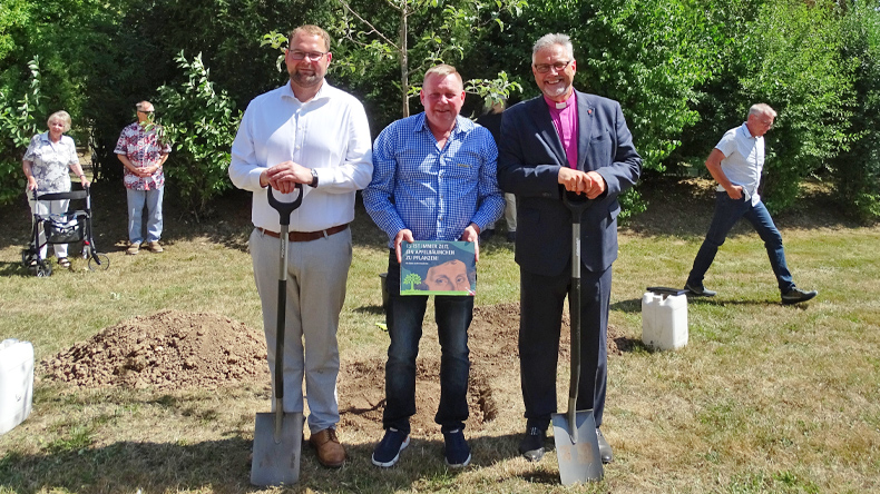Christoph Ihling, Rainer K&ouml;nig und Bischof Werner Philipp stehen vor einem jungen Apfelbaum. Ihling und Philipp halten Spaten, K&ouml;nig zeigt ein Schild zur Baumpflanzung; im Hintergrund sind weitere Teilnehmende zu sehen.