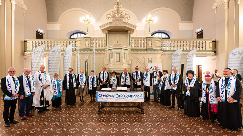 18 kirchenleitende Vertreterinnen und Vertreter unterzeichneten in Berlin die überarbeitete Charta Oecumenica 14 Männer und 4 Frauen stehen im Halbrund um einen Abendmahlstisch mit aufgeschlagenem Buch. Alle Personen tragen ein Tuch mit der Aufschrift »Charta Oecumenica« um den Hals. Im Hintergrund befindet sich eine Kanzel und stehen zwei weitere Personen.