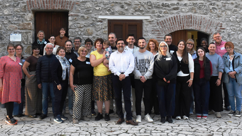 Gruppenfoto: 28 Menschen, davon ein S&auml;ugling auf einem Arm einer Frau, stehen vor einem Haus aus Natursteinen. Frauen sind in der Mehrzahl. Die Personen geh&ouml;ren zu unterschiedlichen Altersgruppen. Manche von ihnen tragen Namensschilder.