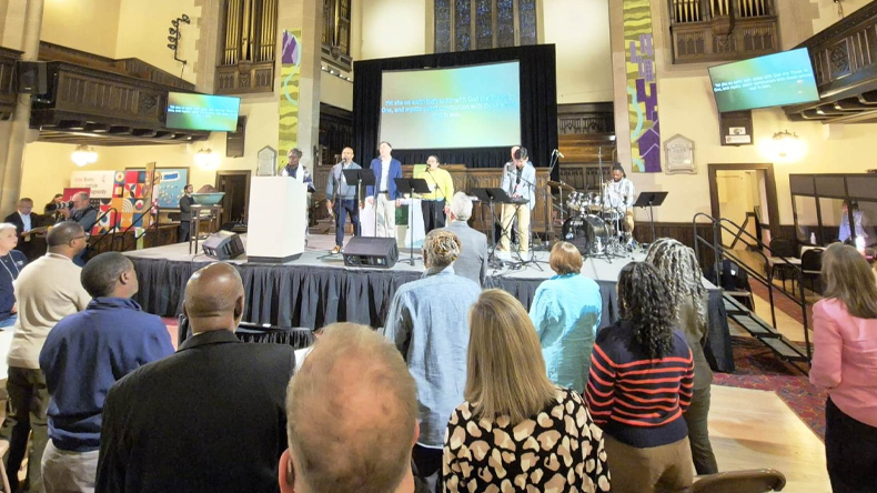 Blick in die Knox United Church in Calgary: Zahlreiche Teilnehmende stehen mit Blick auf die B&uuml;hne. Dort singen und musizieren mehrere Personen. Im Hintergrund sind Orgelpfeifen und hohe Kirchenfenster zu sehen.