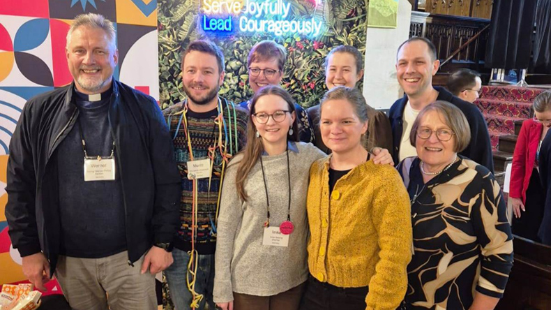 Die deutsche Gruppe in Calgary. Vorn (v. l.): Bischof Werner Philipp, Moritz Mosebach, Imke Sewing, Pastorin Marie-Theres Ringeis, Bischöfin i. R. Rosemarie Wenner. Hinten: Pastorin Anne Detjen, Deborah Holzapfel, Richard Künzel. Acht Teilnehmende aus Deutschland stehen in zwei Reihen für ein Gruppenfoto beim EmK-Leitungstreffen in Calgary. Hinter ihnen sind ein buntes Veranstaltungsmotiv und die Worte »Lead Courageously« zu sehen.