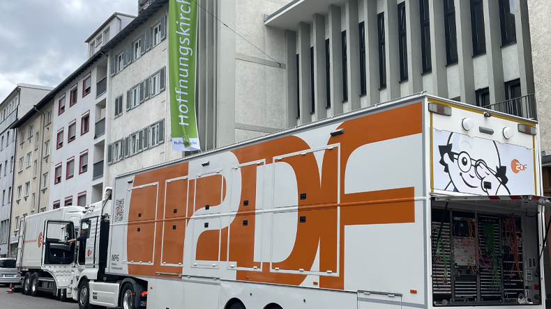 Ein gro&szlig;er wei&szlig;-orangefarbener ZDF-&Uuml;bertragungswagen steht vor der Hoffnungskirche in Stuttgart. Am ge&ouml;ffneten Heck sind zahlreiche technische Ger&auml;te und Kabel zu sehen. Im Hintergrund h&auml;ngt eine gr&uuml;ne Fahne mit der Aufschrift &raquo;Hoffnungskirche&laquo;.